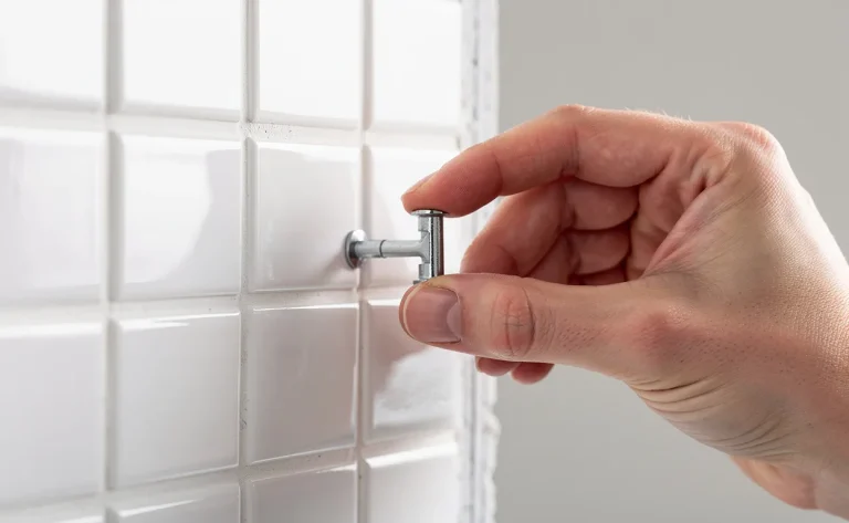 Une main tient une cheville d’ancrage à côté d’une plaque de plâtre blanche recouverte de carreaux blancs lustrés, baignée par une lumière douce.