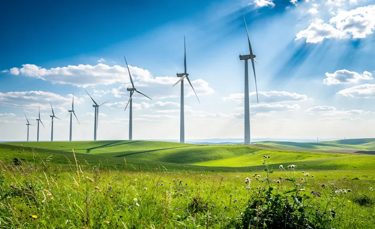 Des éoliennes modernes se dressent dans un champ verdoyant sous un ciel bleu avec quelques rayons de soleil perçant les nuages.
