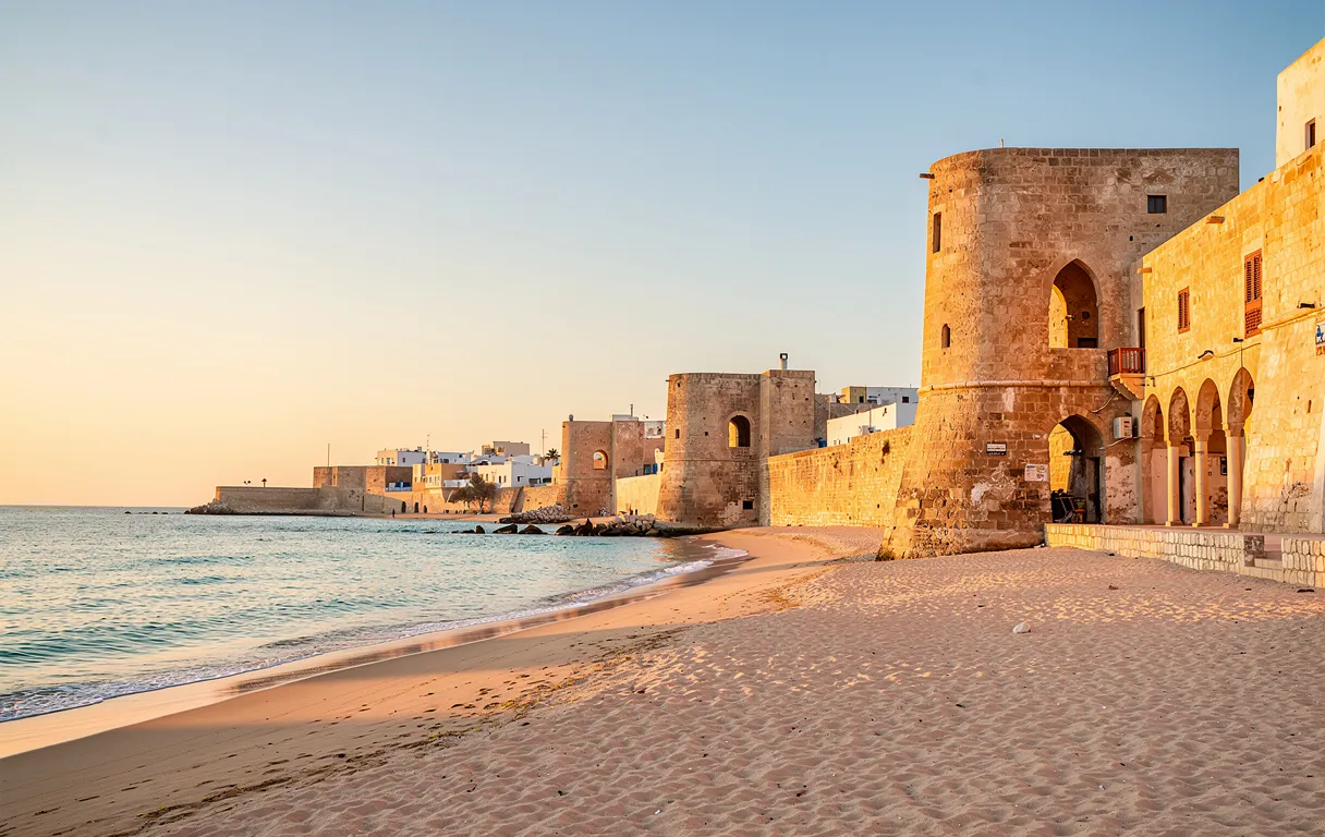 Vue photoréaliste du littoral de Mahdia au coucher du soleil, avec des bâtisses en pierre ancienne devant des eaux turquoise et une plage paisible.